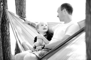 A father and son are sitting in a hammock looking at each other and smiling.