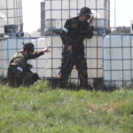 Airsoft players crouch behind a barrier during outdoor airsoft play.